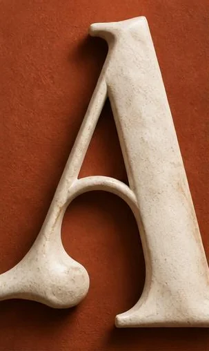 Tegeltableau met letter a in reliëf: een grote witte schreefletter A staat als sculptuur voor een roestkleurige achtergrond.